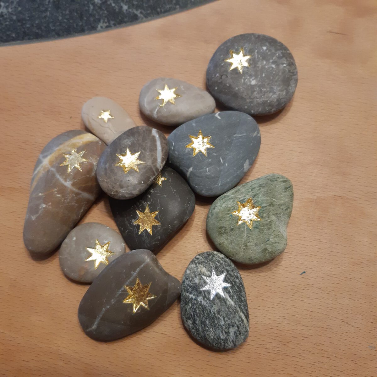 Sternensteine mehrere Kieselsteine liegen auf einer hölzernen Unterlage, die Steinchen sind glatt geschliffen und jeweils mit einem Stern, der vertieft eingearbeitet ist verziert, die Sterne sind gold- oder silberfarbig